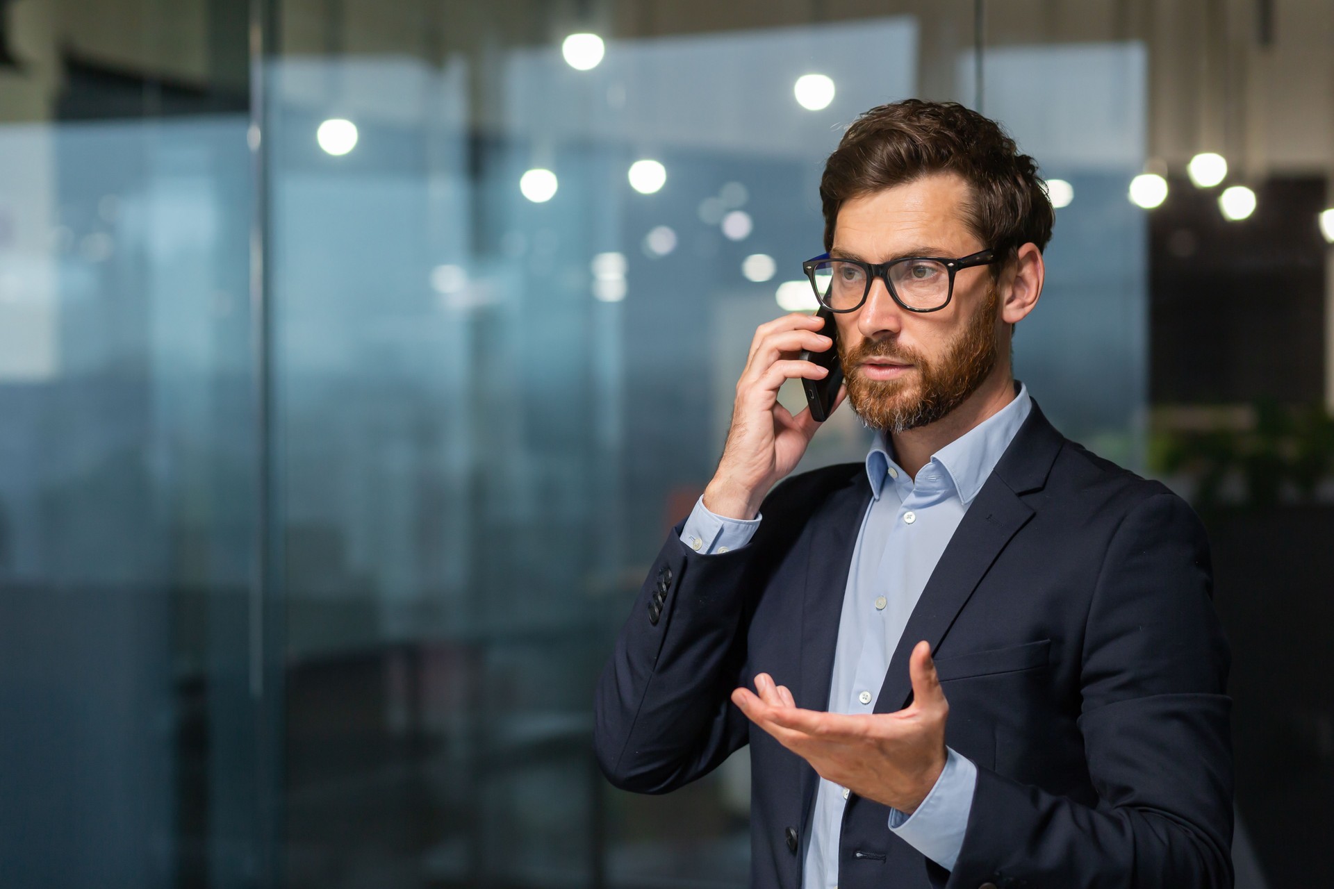 Serious and successful mature businessman inside office near window talking on phone, man in business suit and beard standing